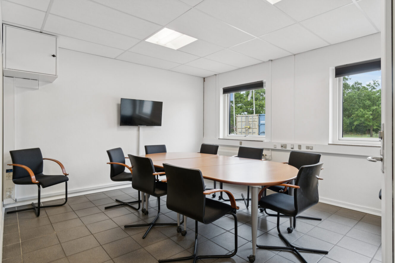 Et lille mødelokale med et rundt bord, syv sorte stole, et væghængt tv, hvide vægge, klinkegulv og to vinduer med grønt udenfor.