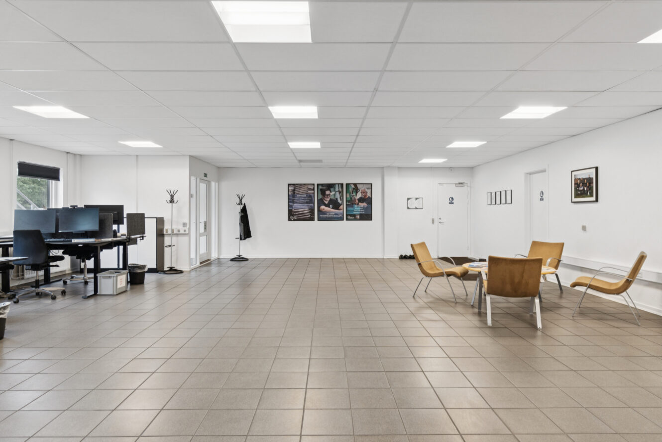 Et rummeligt, moderne kontor med klinkegulv, flere skriveborde med computere til venstre og et lille opholdsområde med fire stole og et rundt bord til højre.