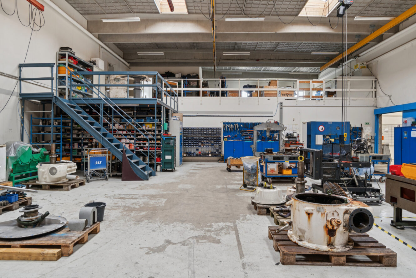 Rummeligt industrielt værksted med hylder, maskindele, værktøj og en metaltrappe; udstyr og materialer er organiseret i hele området.