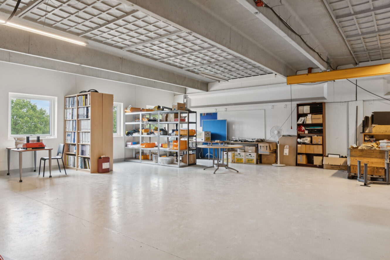 Rummeligt industrielt arbejdsområde med hylder med kasser, skriveborde og kontorartikler, betongulve og store vinduer, der lukker naturligt lys ind.