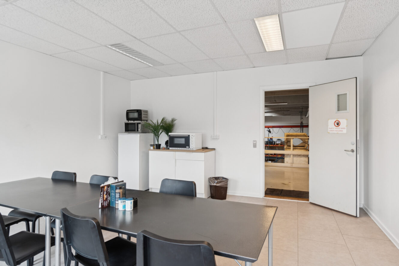 Et pauserum med et sort bord og flere stole, et køleskab, to mikroovne, en kaffemaskine og en åben dør, der fører til et værkstedsområde.