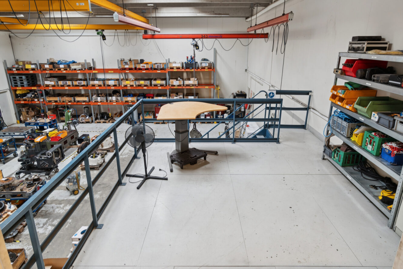 Et industrielt værksted med metalhylder med værktøj og udstyr, set fra en balkon på øverste etage med et stående skrivebord og en ventilator.