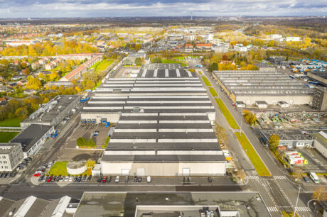 Luftfoto af et stort industrielt lagerkompleks omgivet af veje, parkeringspladser og nærliggende bygninger i et byområde.