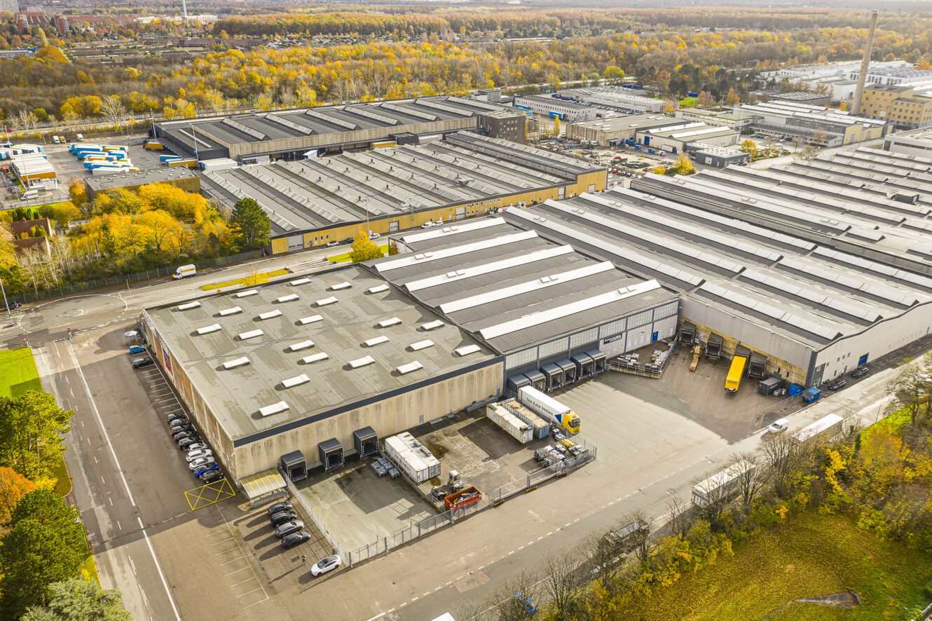 Luftfoto af et stort industrikompleks med flere lagerbygninger, læsseramper, lastbiler og omkringliggende veje; efterårstræer ses i baggrunden.