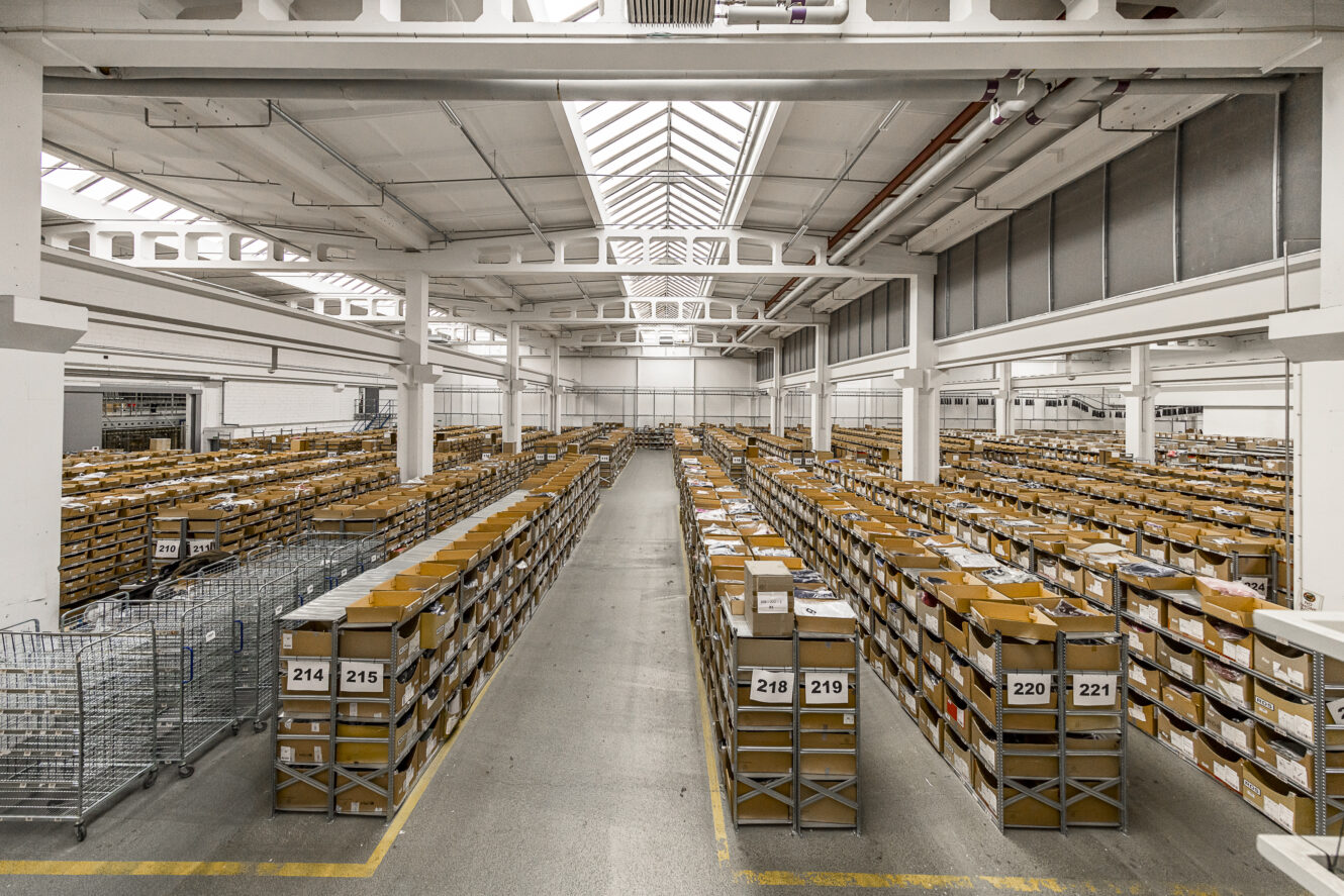 Stort lagerinteriør med rækker af metalhylder fyldt med mærkede papkasser, oplyst af ovenlysvinduer og loftslamper.