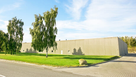 Stor, vinduesløs beige bygning med fladt tag, omgivet af grønt græs, et par træer og en asfalteret indkørsel ved siden af en vej under en delvis overskyet himmel.