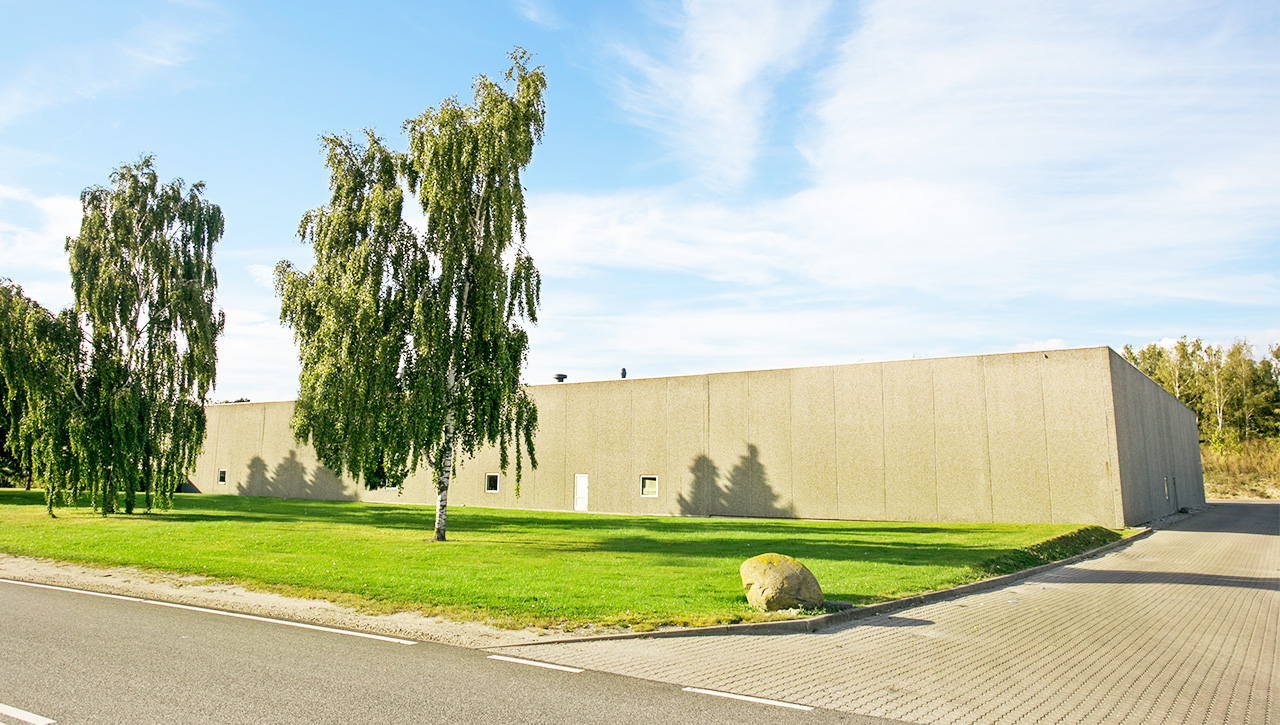 Stor, vinduesløs beige bygning med fladt tag, omgivet af grønt græs, et par træer og en asfalteret indkørsel ved siden af en vej under en delvis overskyet himmel.