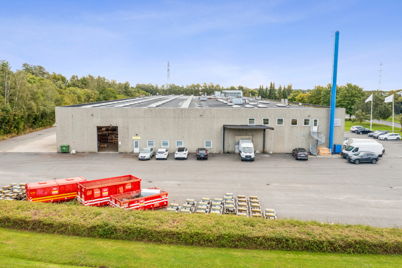 Et stort industrilager med et gråt ydre, flere parkerede køretøjer foran, røde affaldscontainere og stabler af materialer på den asfalterede grund. Træer og græs omgiver ejendommen.