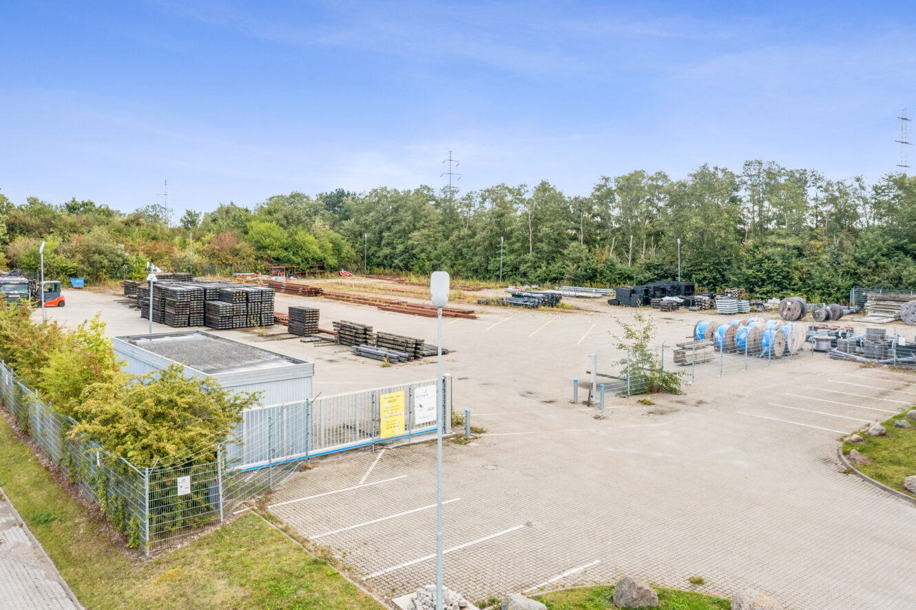 En industriel lagerplads med stablede metalmaterialer, hegn, lysmaster og træer i baggrunden under en klar blå himmel.