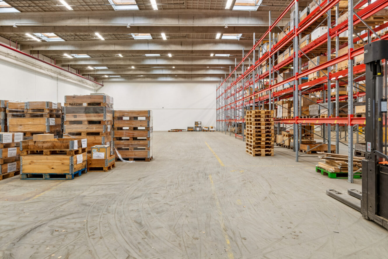 Rummelig lagerbygning med højt til loftet, tomme røde metalreoler, stablede træpaller og spredte kasser på betongulvet.