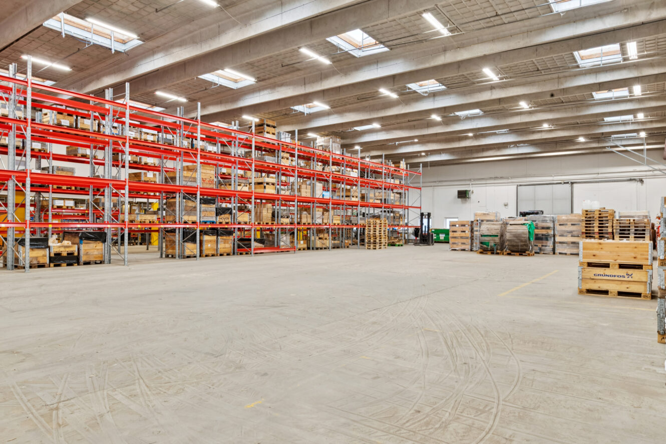 Stort lagerinteriør med høje metalhylder med trækasser og paller, stort tomt gulv og ovenlys.