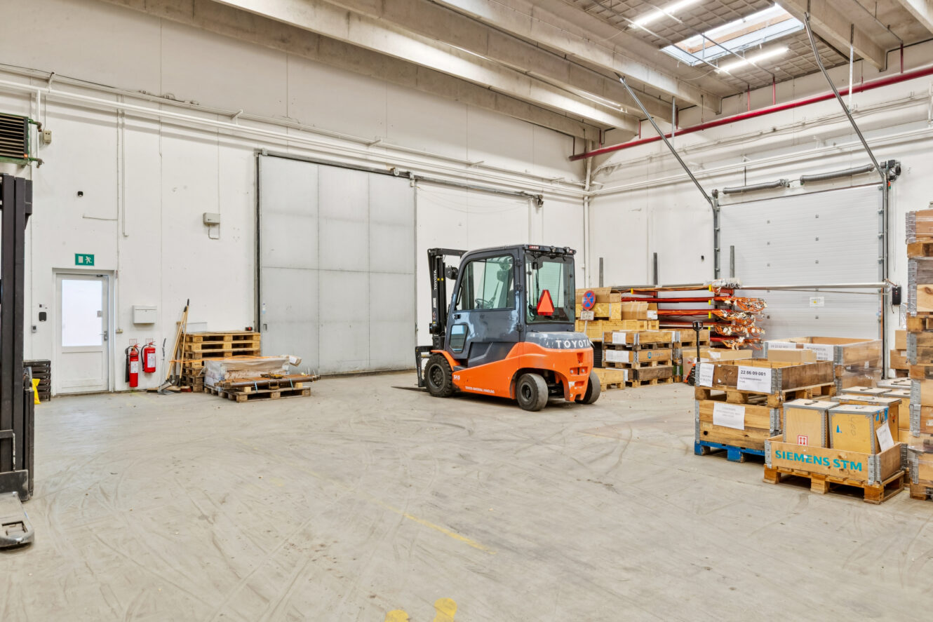 En orange gaffeltruck er parkeret i en rummelig lagerhal, omgivet af træpaller og stablede papkasser.
