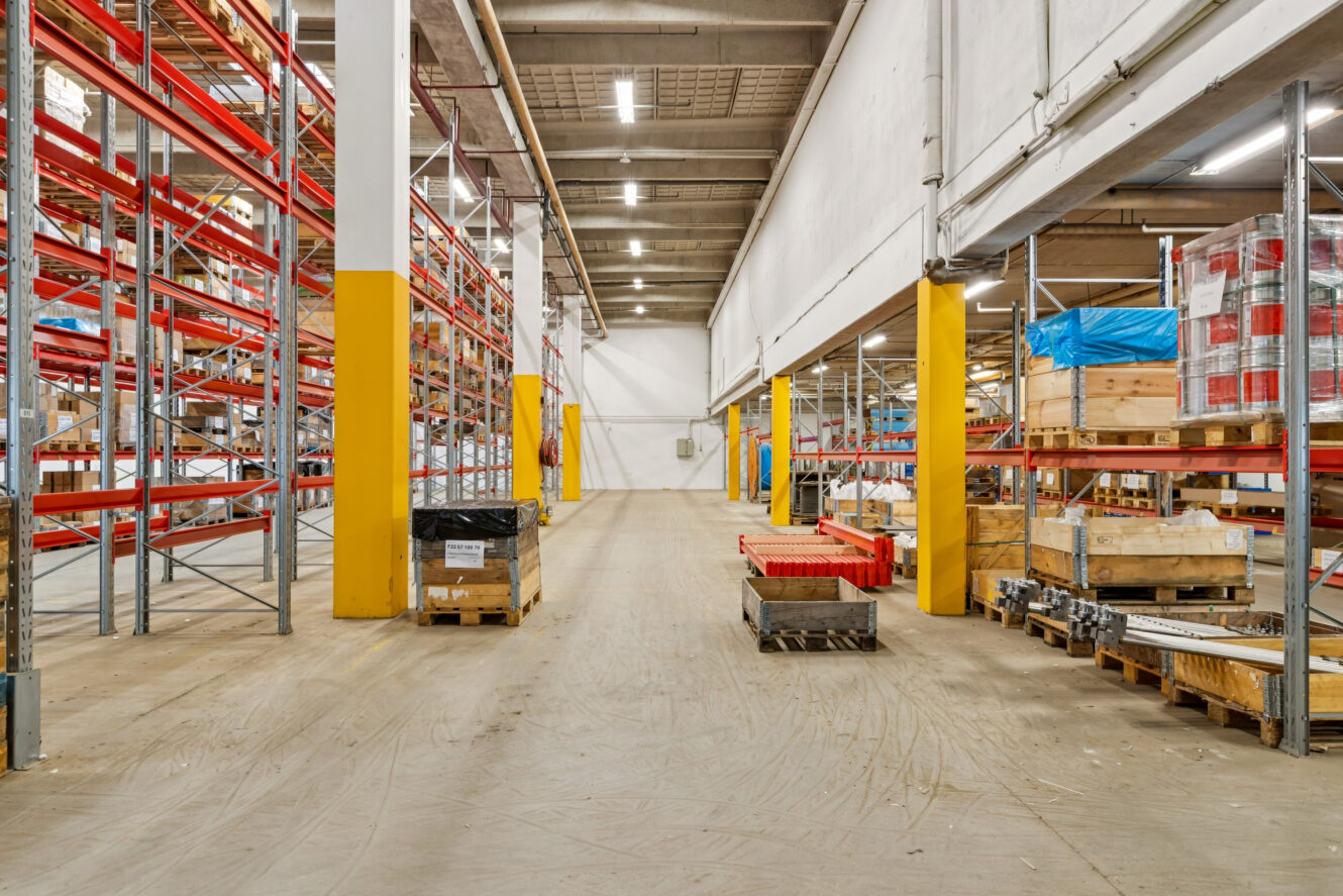 En rummelig lagerbygning med tomme røde metalreoler, gule søjler, træpaller og diverse pakkede varer på gulvet.