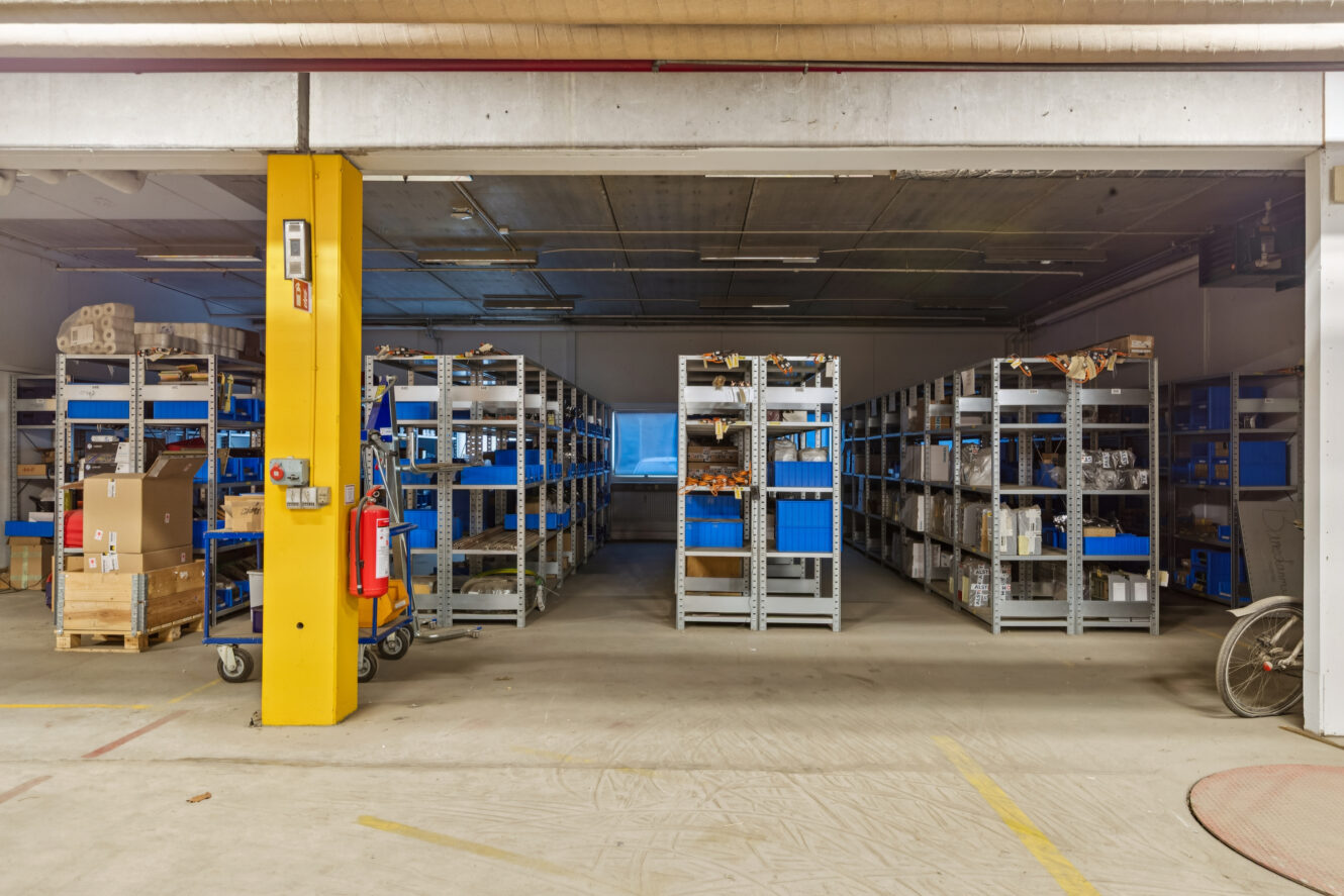Indendørs lagerbygning med metalhylder i rækker med forskellige kasser og beholdere; en vogn og en brandslukker står nær en gul konstruktionssøjle i forgrunden.
