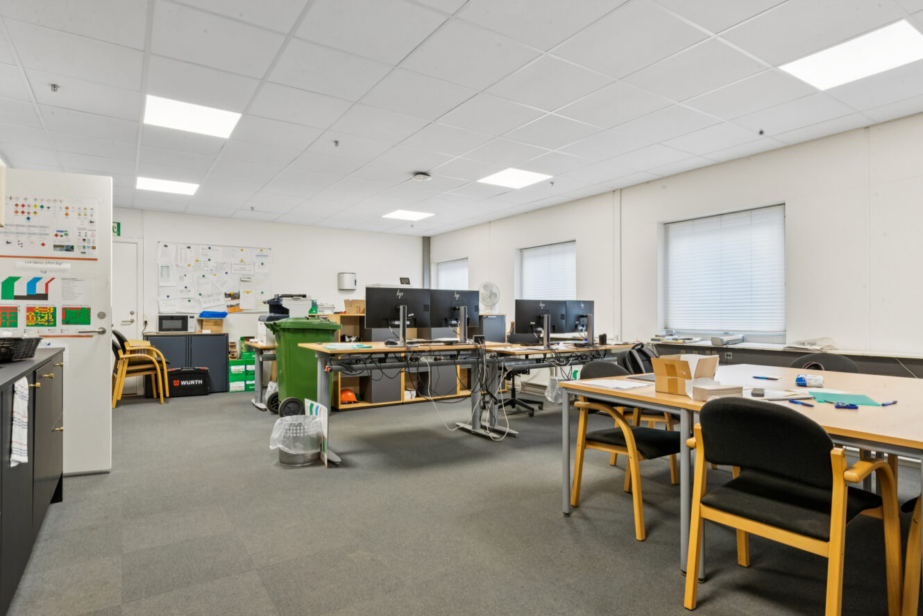 Kontorlokaler med flere skriveborde, computerskærme, stole og opslagstavler på væggene; vinduer lukker naturligt lys ind.