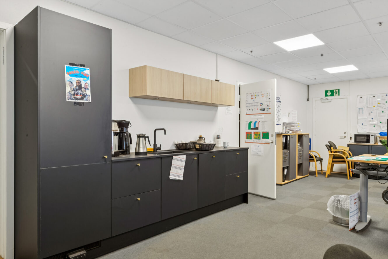 Pauserum på kontoret med et sort tekøkken, kaffemaskine, vandkedel, træskabe og forskellige papirer og plakater på væggene. Gule stole og et bord ses i baggrunden.