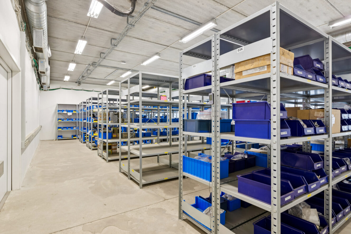 Industrielt lagerrum med metalhylder med blå plastkasser og papkasser, organiseret i rækker under fluorescerende belysning. Gulvet er af beton, og loftet har synlige kanaler.
