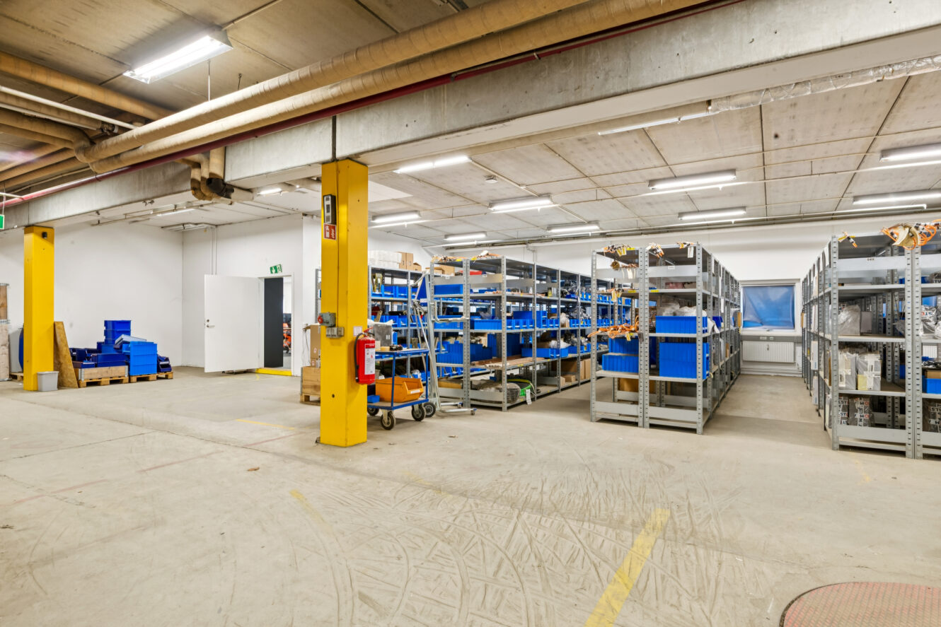 Industrielt lagerinteriør med metalreoler med blå beholdere, forskelligt udstyr og forsyninger; gule søjler og betongulv er synlige.