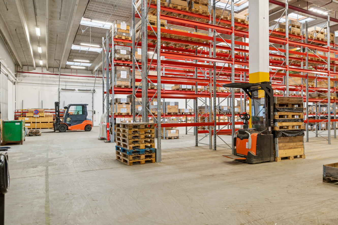 Et lagerinteriør med høje røde hylder med kasser, træpaller stablet på gulvet og to gaffeltrucks parkeret på åbne pladser.