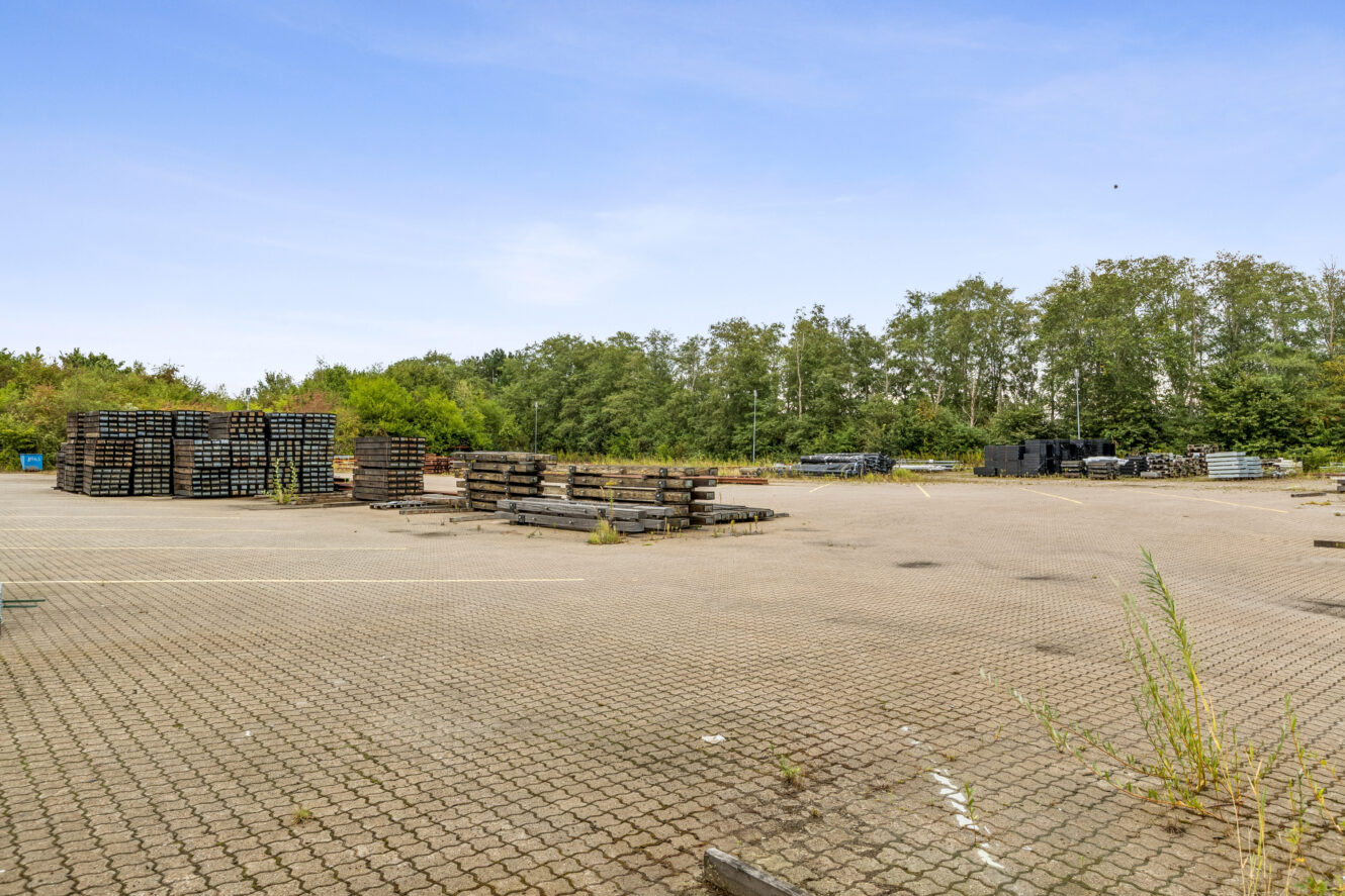 Stabler af metalrør og bjælker er anbragt på en stor asfalteret udendørs plads omgivet af træer under en klar himmel.