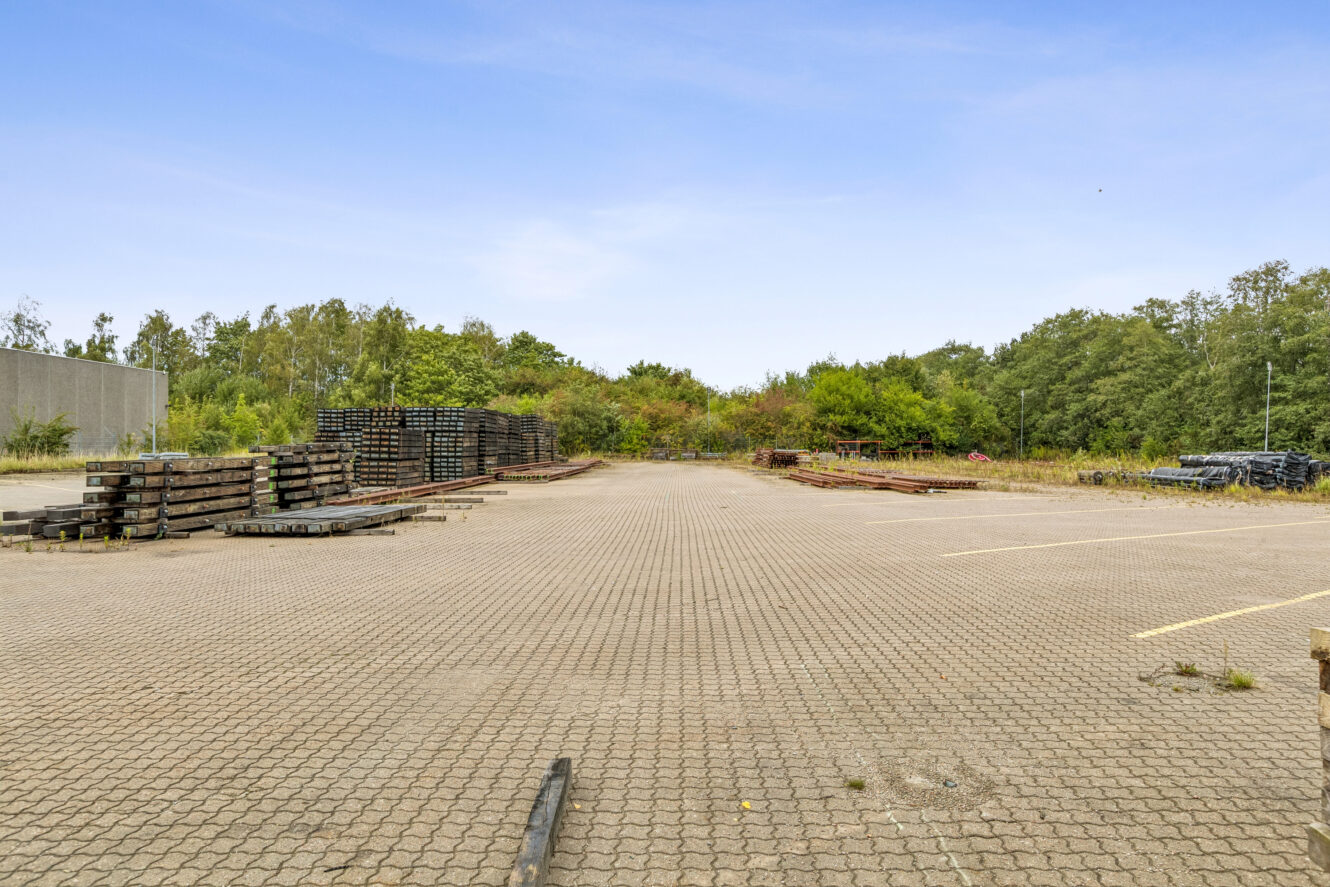 En tom asfalteret grund med stabler af træbjælker og træer i baggrunden under en klar himmel.
