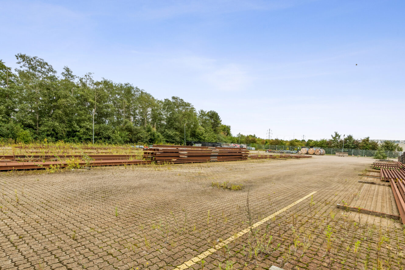 Udendørs lagerplads med stabler af metalbjælker på en asfalteret overflade, omgivet af træer og vegetation under en klar himmel.