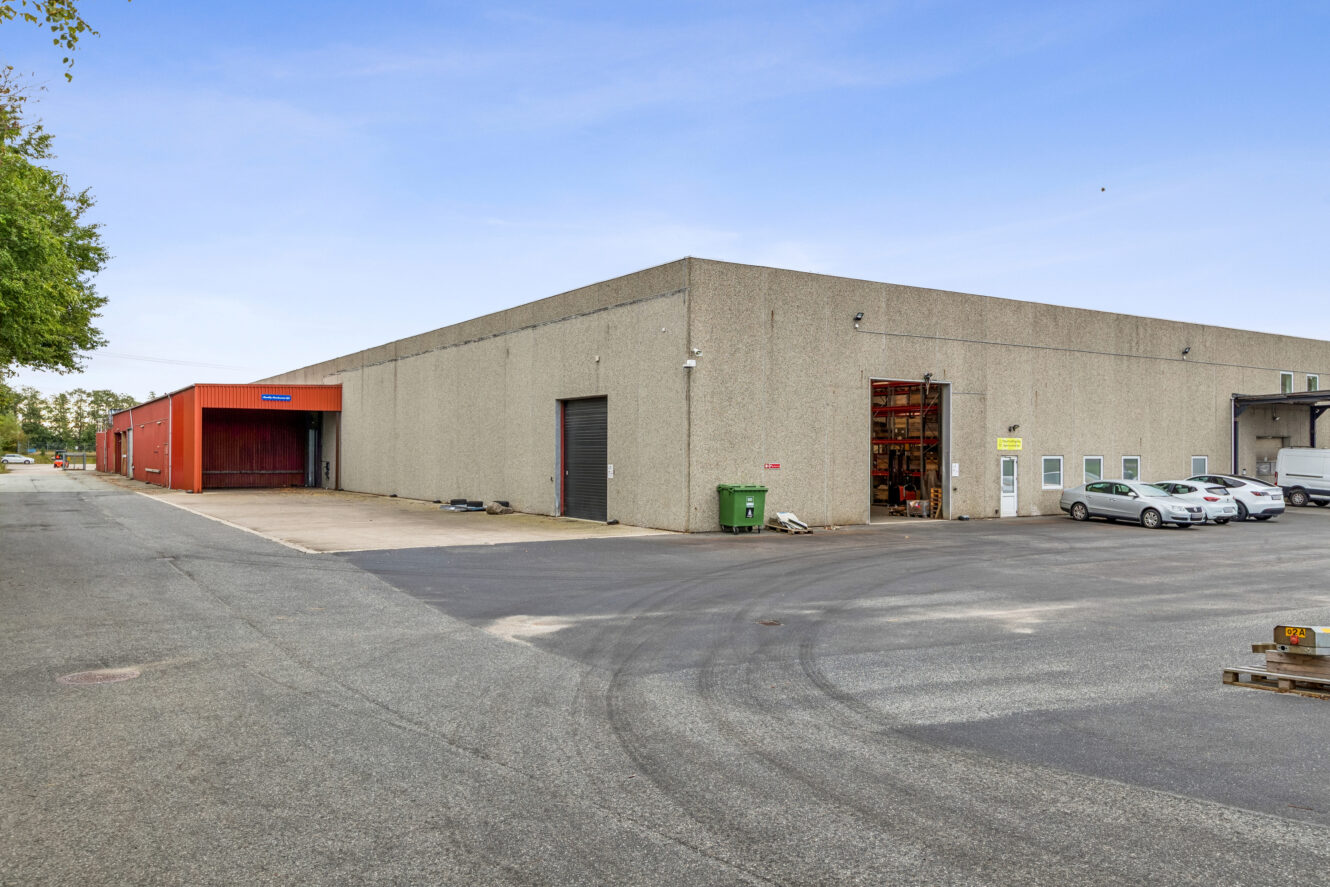 Stort industrilager med solbrændt beton udvendigt, læsseramper, flere parkerede biler og et rødt indgangsparti på en asfalteret grund under en klar himmel.