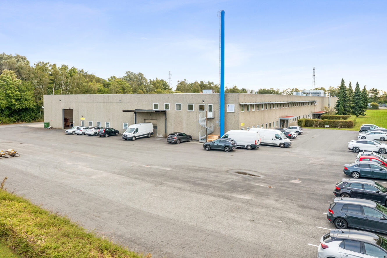 Stort industrilager med flere læsseramper, parkerede varevogne og biler foran, omgivet af træer og åben plads under en klar himmel.