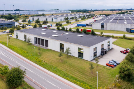 Et moderne industrilager med fladt tag og store vinduer, omgivet af græs og parkeringspladser, beliggende i en erhvervspark med lignende bygninger.