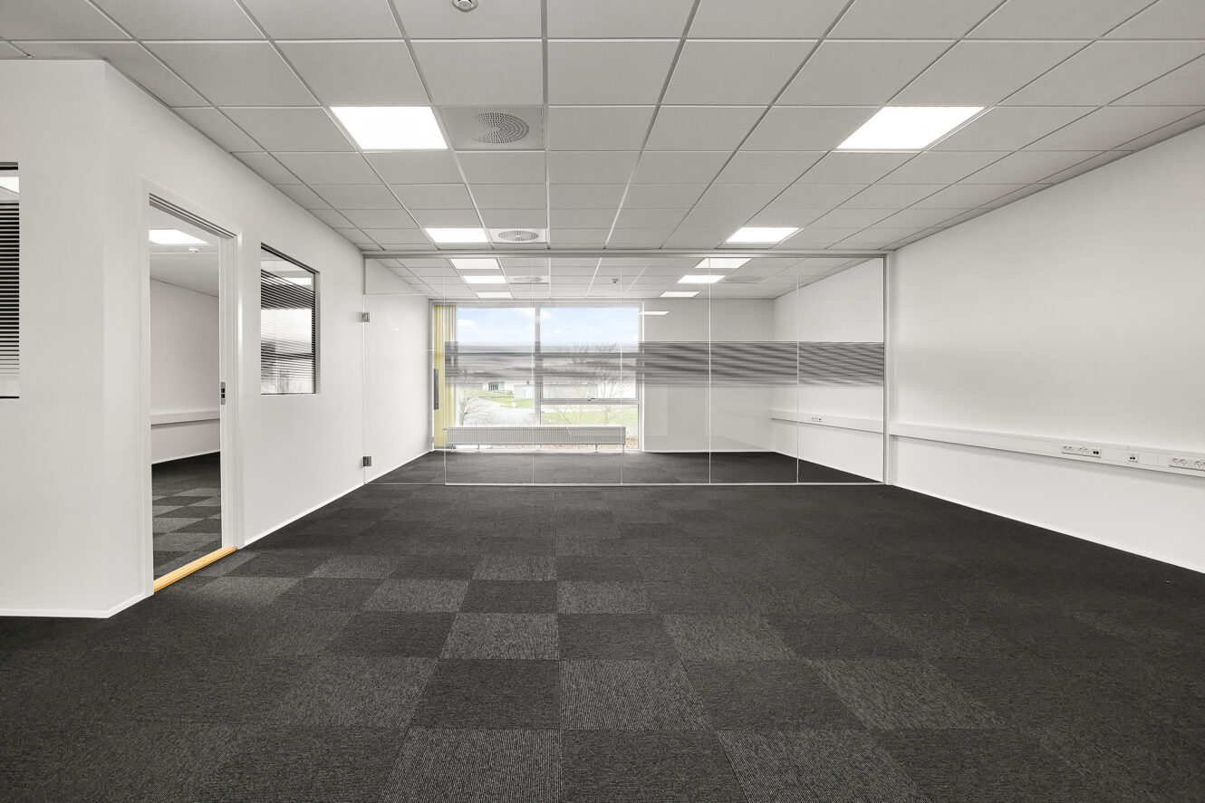 Tomt, moderne kontorlokale med gråt tæppe, hvide vægge, loftslamper og store glasvinduer med udsigt til en udendørs scene.