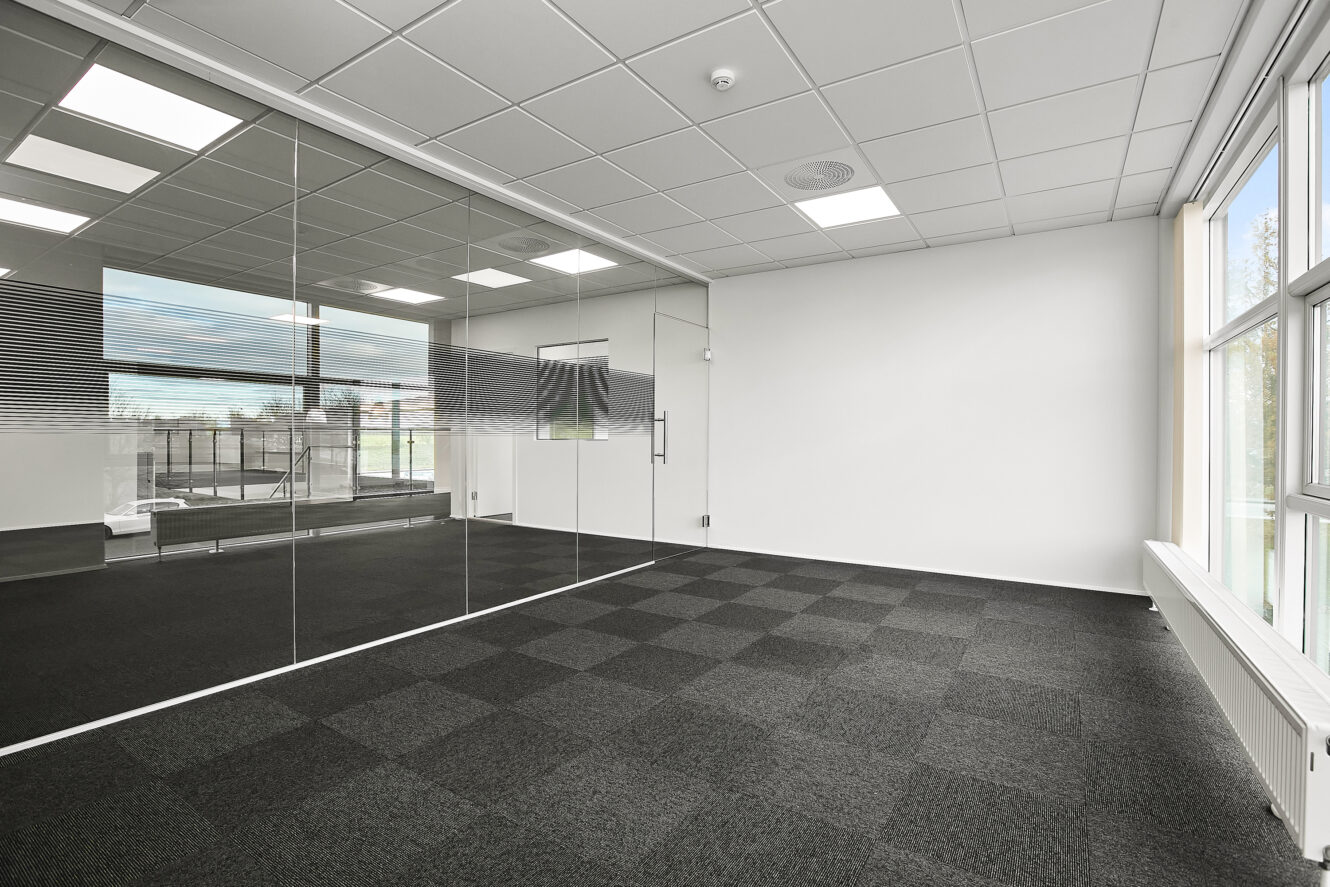 Tomme, moderne kontorlokaler med glasvægge, ternet gulvtæppe, hvide vægge og store vinduer, der lukker naturligt lys ind.