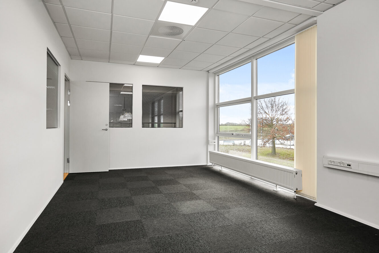 Tomt kontorlokale med hvide vægge, sort gulvtæppe, store vinduer, lysstofrør i loftet og en lukket dør med indvendige vinduer.