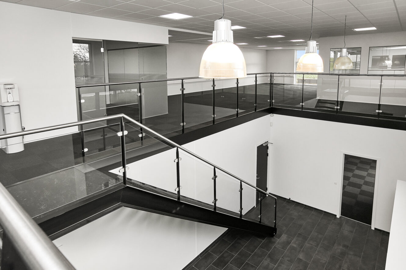 Moderne kontorindretning med en trappe med glasgelænder, store pendellamper og en blanding af kontorer med glasvægge og lukkede døre på begge etager.