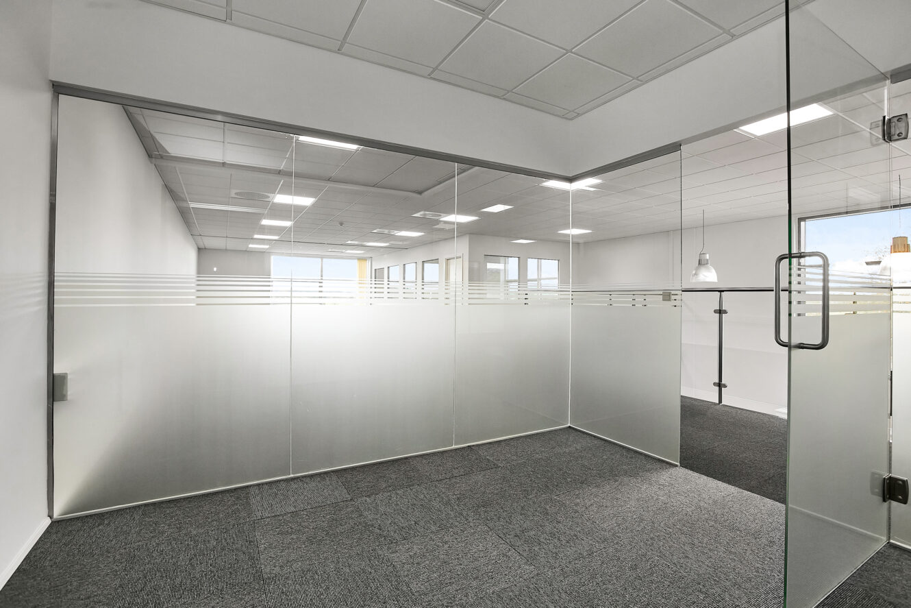 Tomme, moderne kontorlokaler med glasvægge, gråt gulvtæppe, hvide loftsplader og fluorescerende belysning.