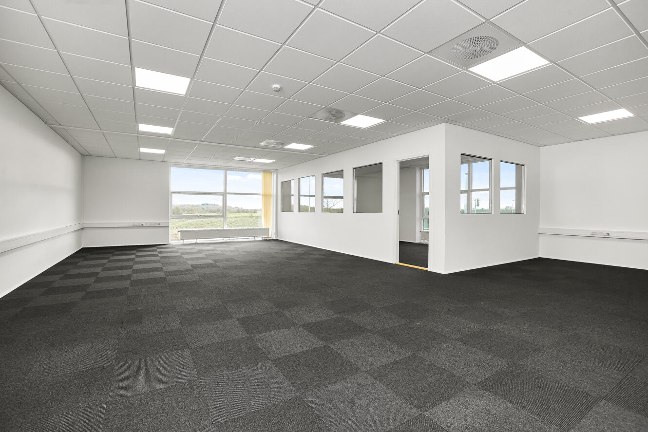Tomme kontorlokaler med hvide vægge, sorte tæppefliser, et mødelokale med glasvægge og store vinduer, der lukker naturligt lys ind.
