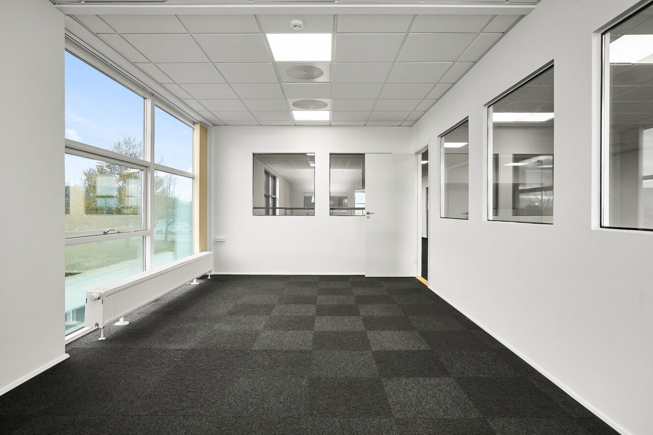 Tomme, moderne kontorlokaler med store vinduer, indvendige glasvægge, en hvid dør og ternet, mørkt gulvtæppe.