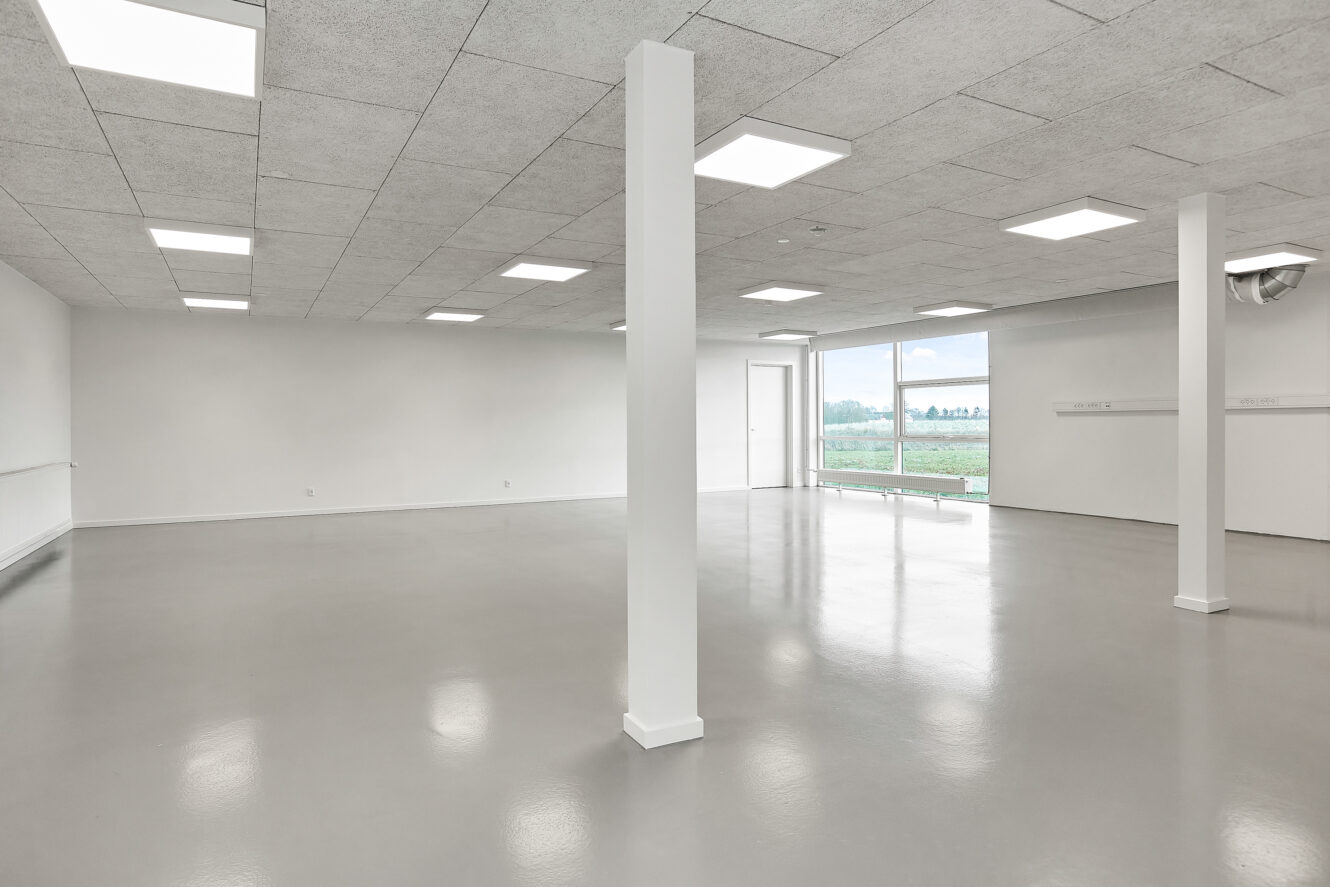 Tomt, moderne rum med hvide vægge, loftplader og store vinduer, der lukker naturligt lys ind. Gulvet er glat og reflekterende, og bærende søjler er fordelt i hele rummet.