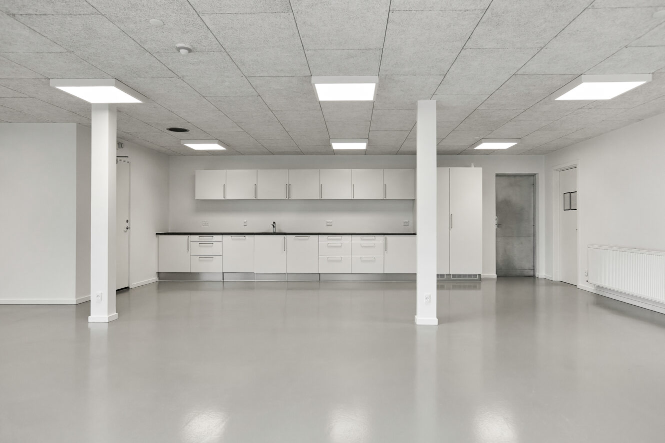 Stort, tomt rum med et minimalistisk hvidt tekøkken, gråt gulv, hvide vægge, to søjler og et loft med indbygget belysning.