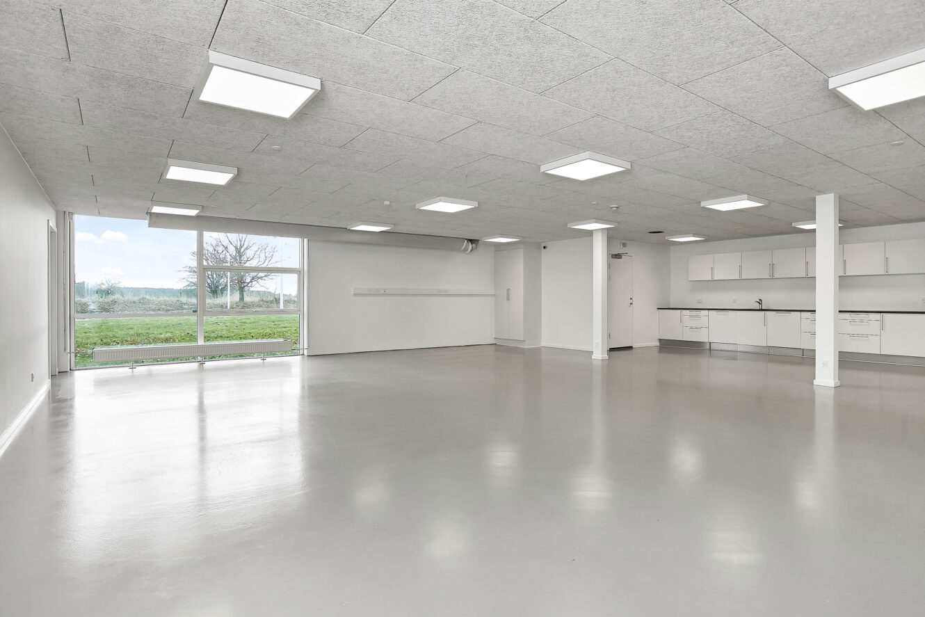 Rummeligt, tomt rum med hvide vægge, store vinduer, loftslamper og et tekøkken langs den ene væg; poleret gråt gulv og udsigt til grønne områder udenfor.