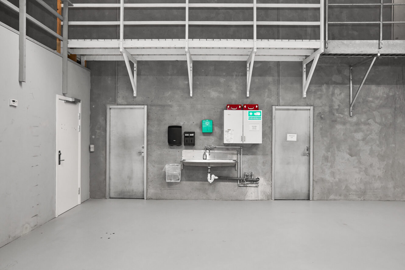 Industriel vaskestation med vask i rustfrit stål, sæbe- og håndklædedispensere, nødforsyninger og to døre op ad en grå betonvæg i en lagerbygning.