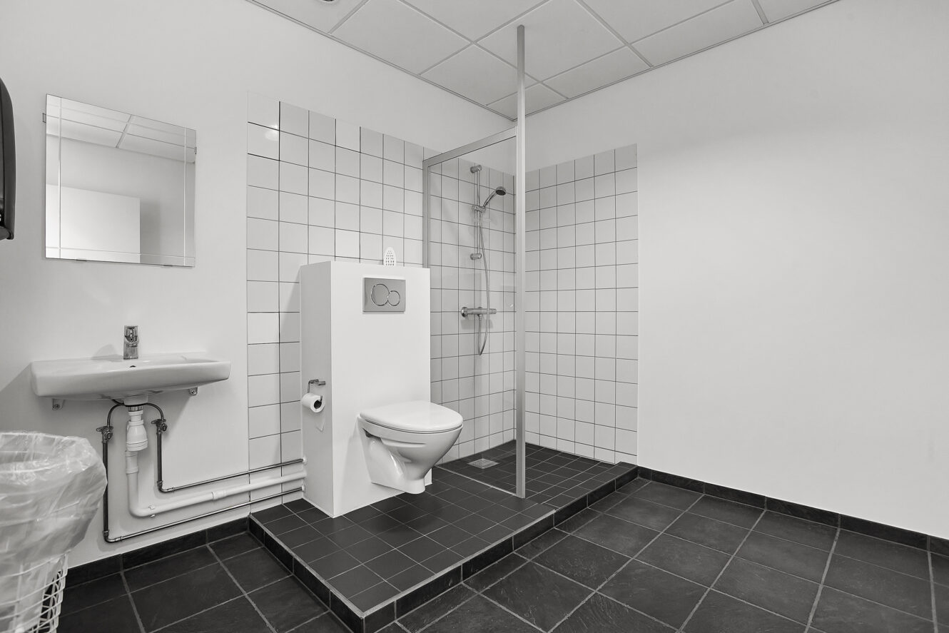 Sort-hvidt foto af et toilet med et væghængt toilet, en håndvask med spejl, en skraldespand og et åbent bruseområde, alt sammen på et flisegulv.