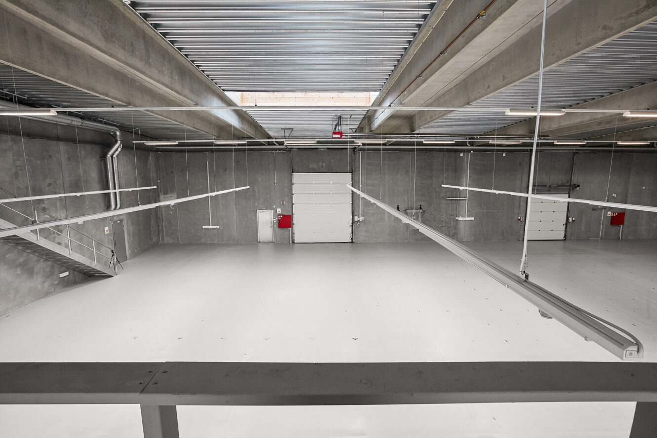 Tomt, moderne industrilager med betonvægge, højt til loftet, stærk belysning, store hvide døre og en metaltrappe i venstre side.