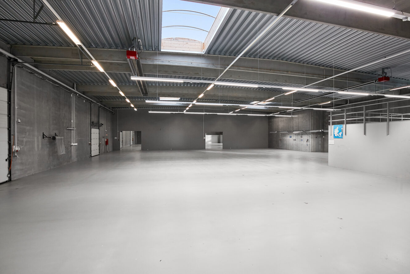 Bredt udsyn over et tomt industrilager med betongulv, højt til loftet, ovenlys og minimalt inventar langs væggene.