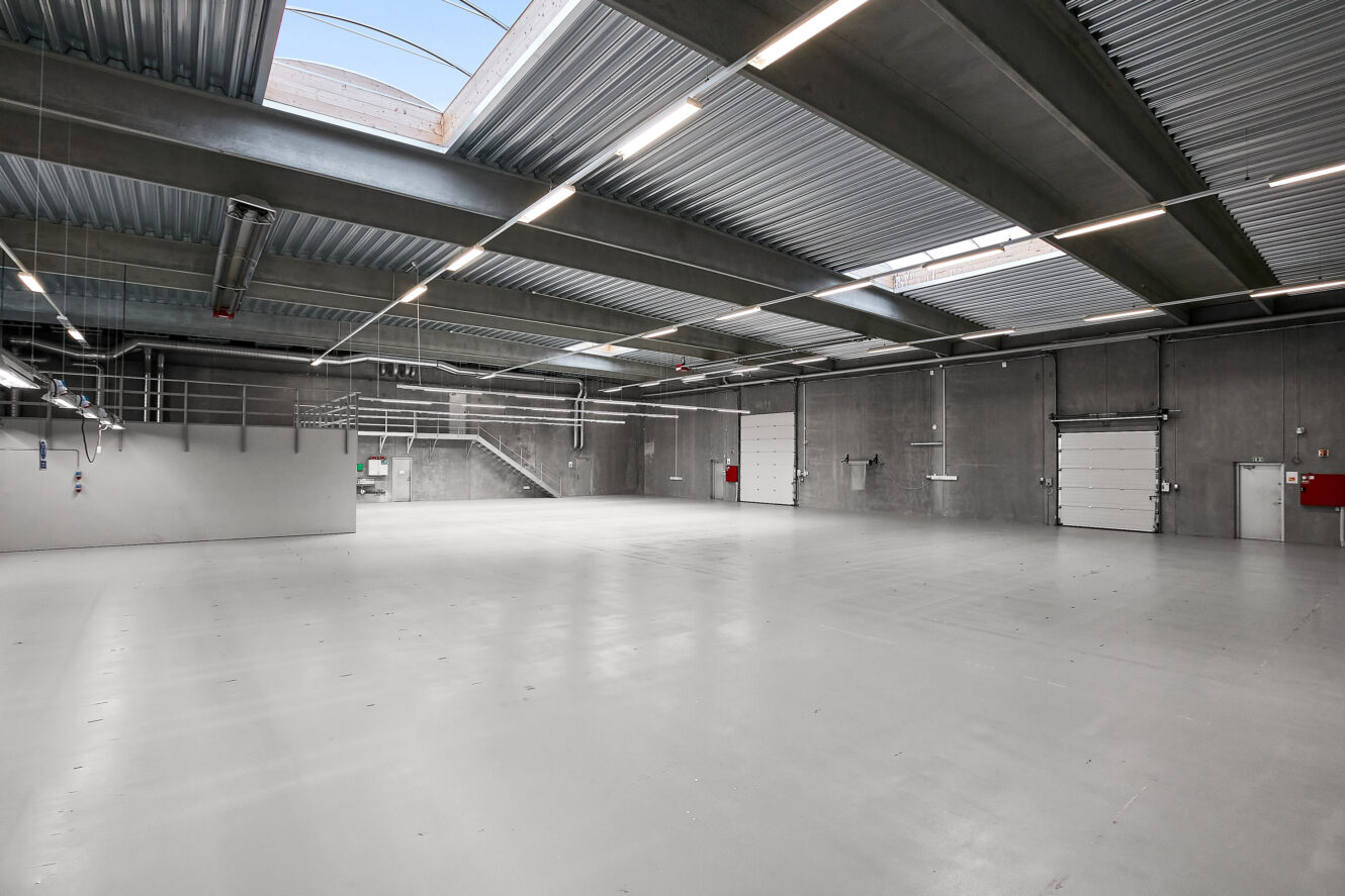 Tomt industrielt lagerinteriør med betongulve, højt til loftet, ovenlys, overlys, store garageporte og synlige konstruktionselementer.