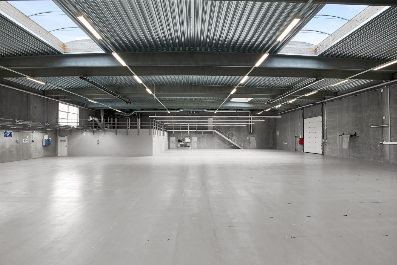 Stort, tomt industrilager med betongulve, højt til loftet, ovenlysvinduer og store ledhejseporte.