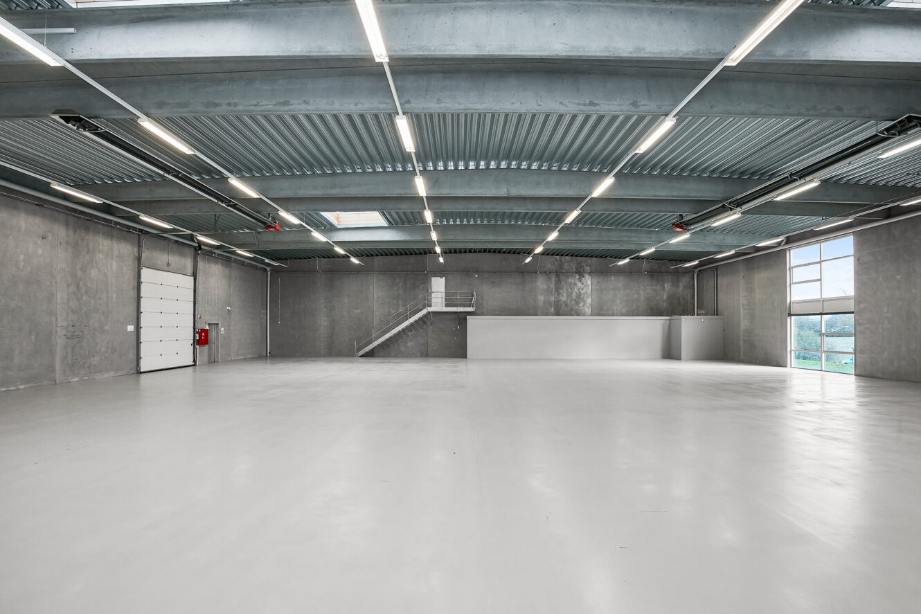 Stort, tomt industrilager med betonvægge og -gulv, lysstofrør, en trappe, store vinduer og en hvid sektionsopdelt garageport.