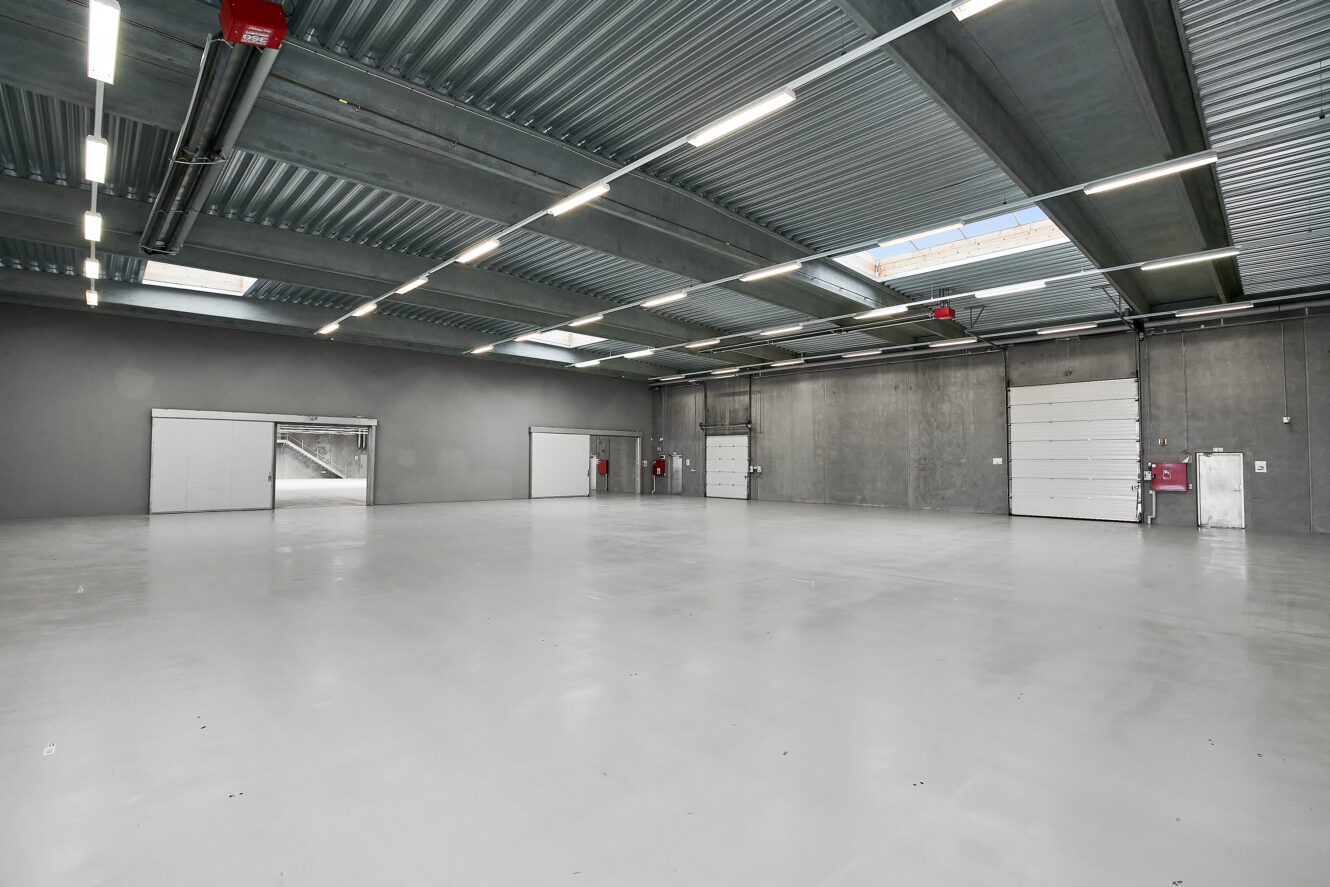 Rummelig, tom lagerbygning med betongulv, fritlagt loft, ovenlys og flere store døre langs væggene.