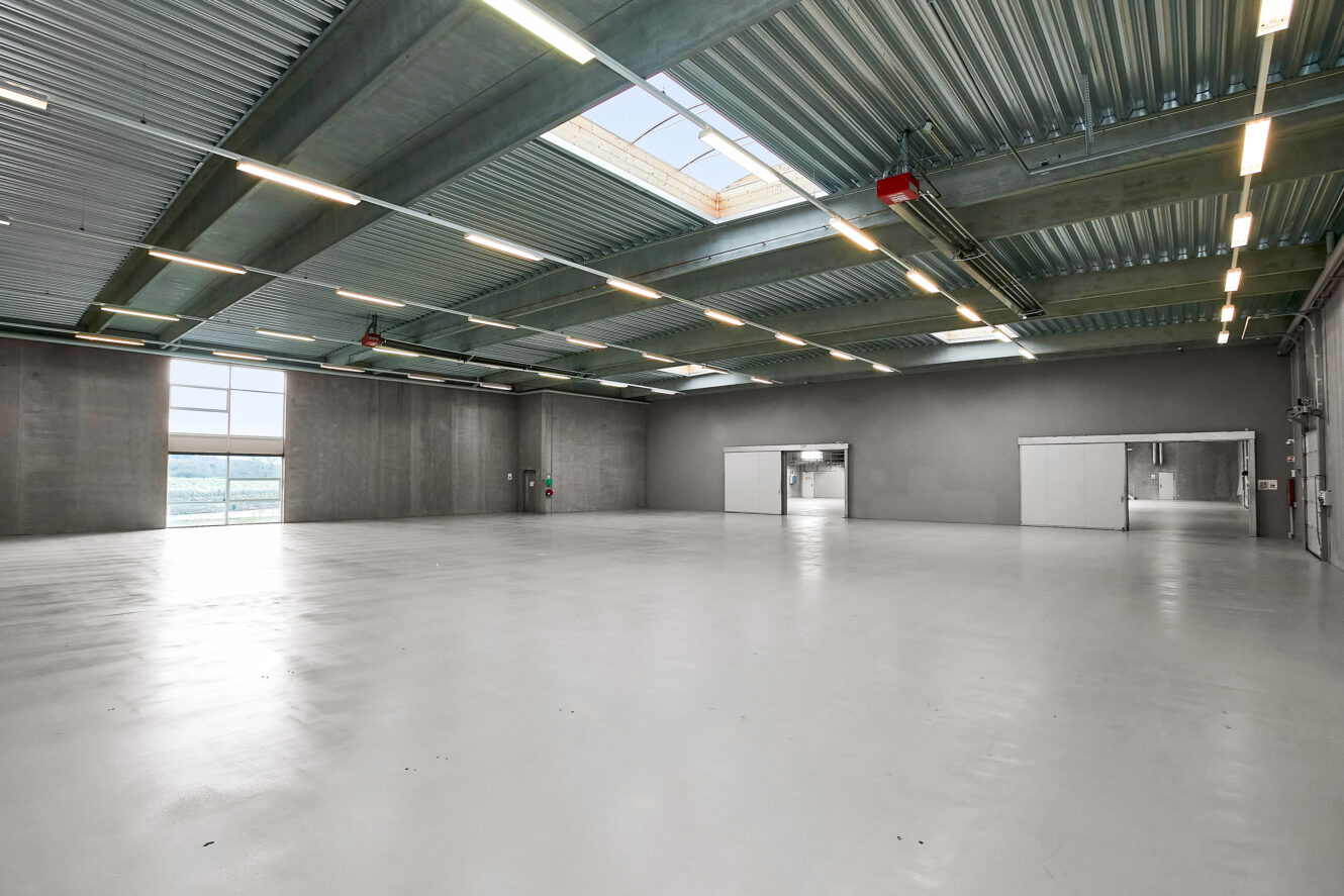 Stort, tomt industrilager med poleret betongulv, højt til loftet, synlige bjælker, ovenlys og store vinduer, der lukker naturligt lys ind.