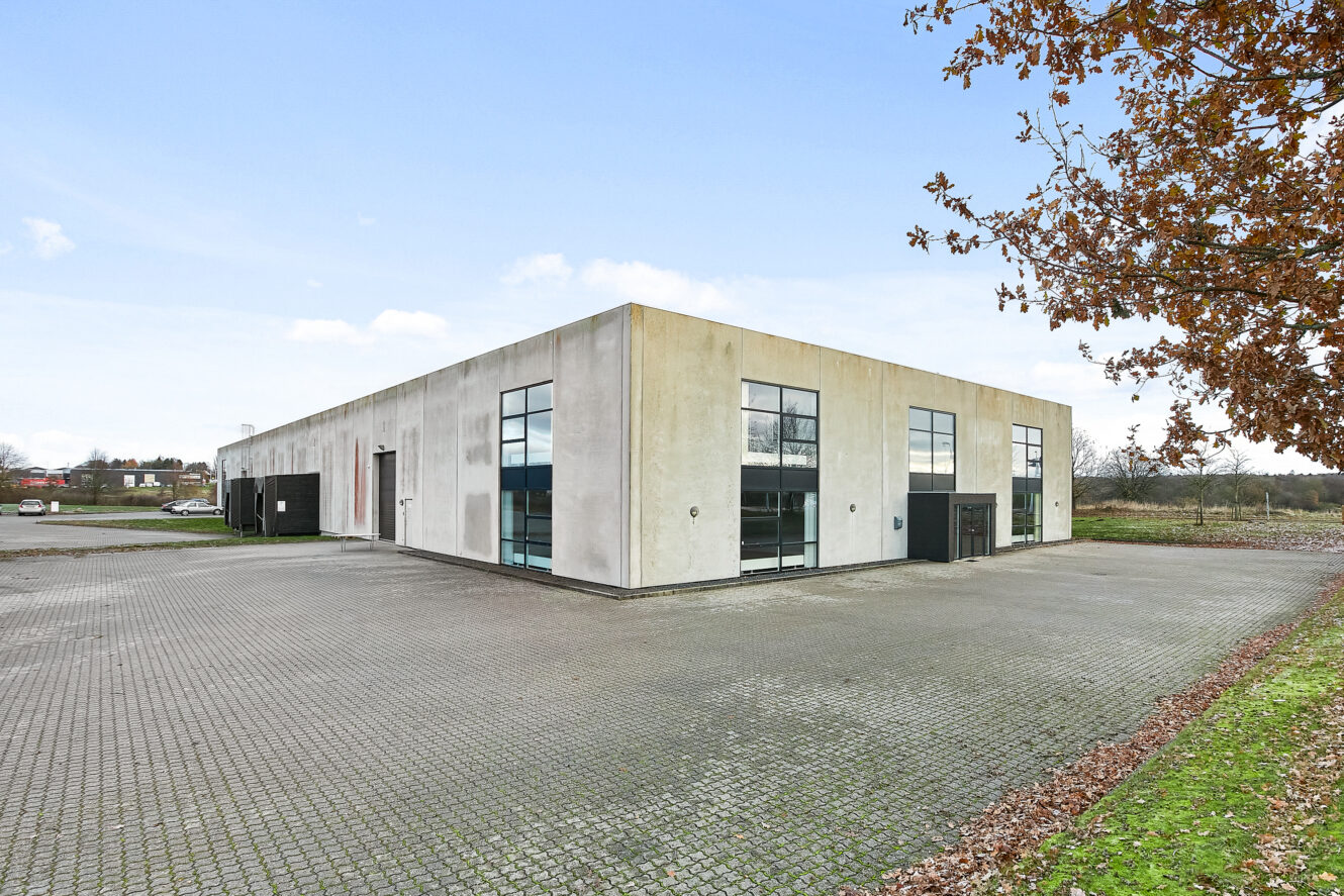 En stor, rektangulær industribygning med flere vinduer og en asfalteret grund, omgivet af sparsomme træer og åbent land under en klar himmel.