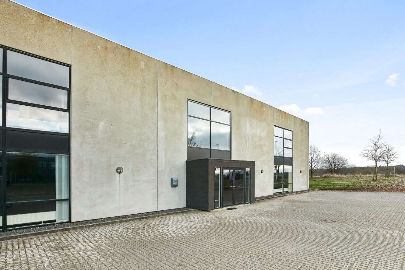 En moderne, rektangulær industribygning med store vinduer og en lille glasindgang, omgivet af et asfalteret område og nogle træer i baggrunden.